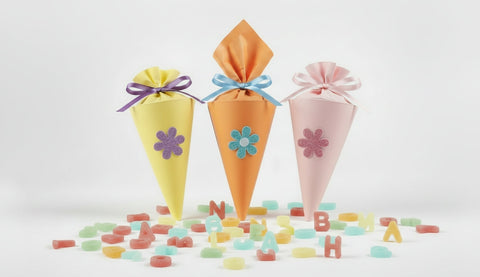 Drei Mini-Schultüten in den Farben gelb, orange und rosa inmitten von bunten Bonbon Buchstaben aus XYLIT. Das Bild ist mit KI erstellt.