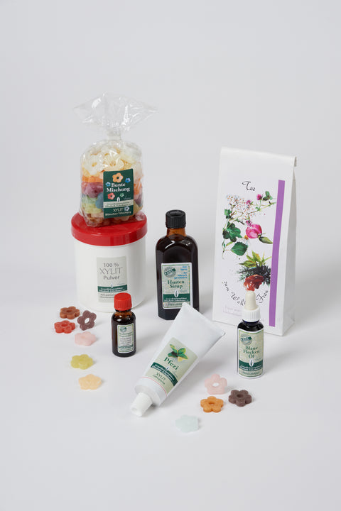 Einzelne Blümchen liegen versträut vor einer Dose Xylit Pulver, ein Beutel XYLIT-BLÜMCHEN Bunte Mischung steht auf der Dose, nebenan eine kleine Flasche Hustensirup, vor der Dose steht eine Flasche Katherogast, daneben liegt eine Tube mit Zahnpasta Pfezi, dahinter steht eine Packung Tee zum Wohl der Frau und davor steht eine Flasche Blaue Flecken Öl auch hier liegen vereinzelt Blümchen zur Deko, Produktwerbebild